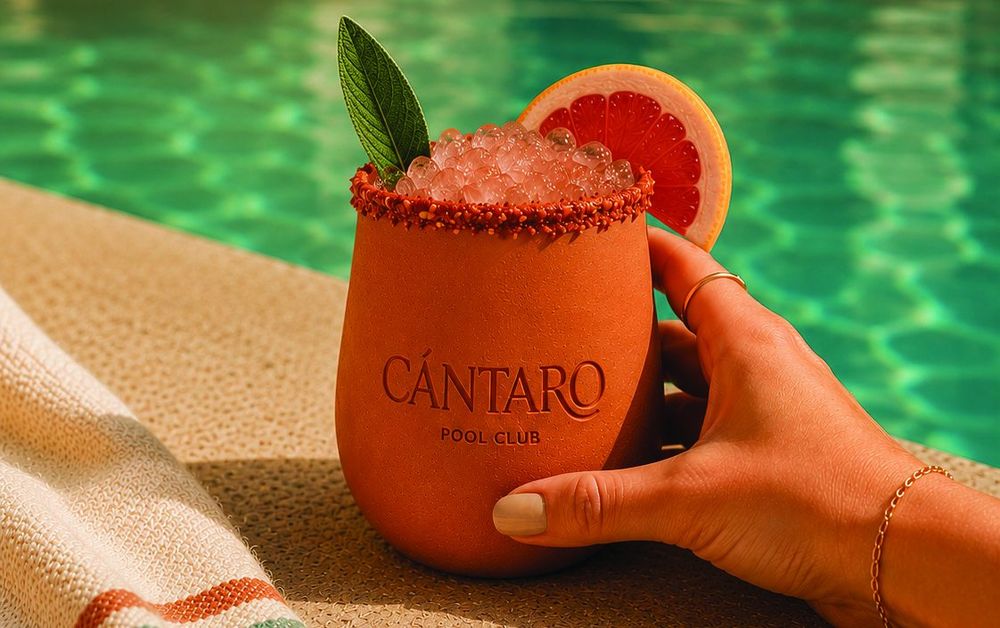 A hand holding a Cántaro clay cup garnished with grapefruit at the edge of the pool