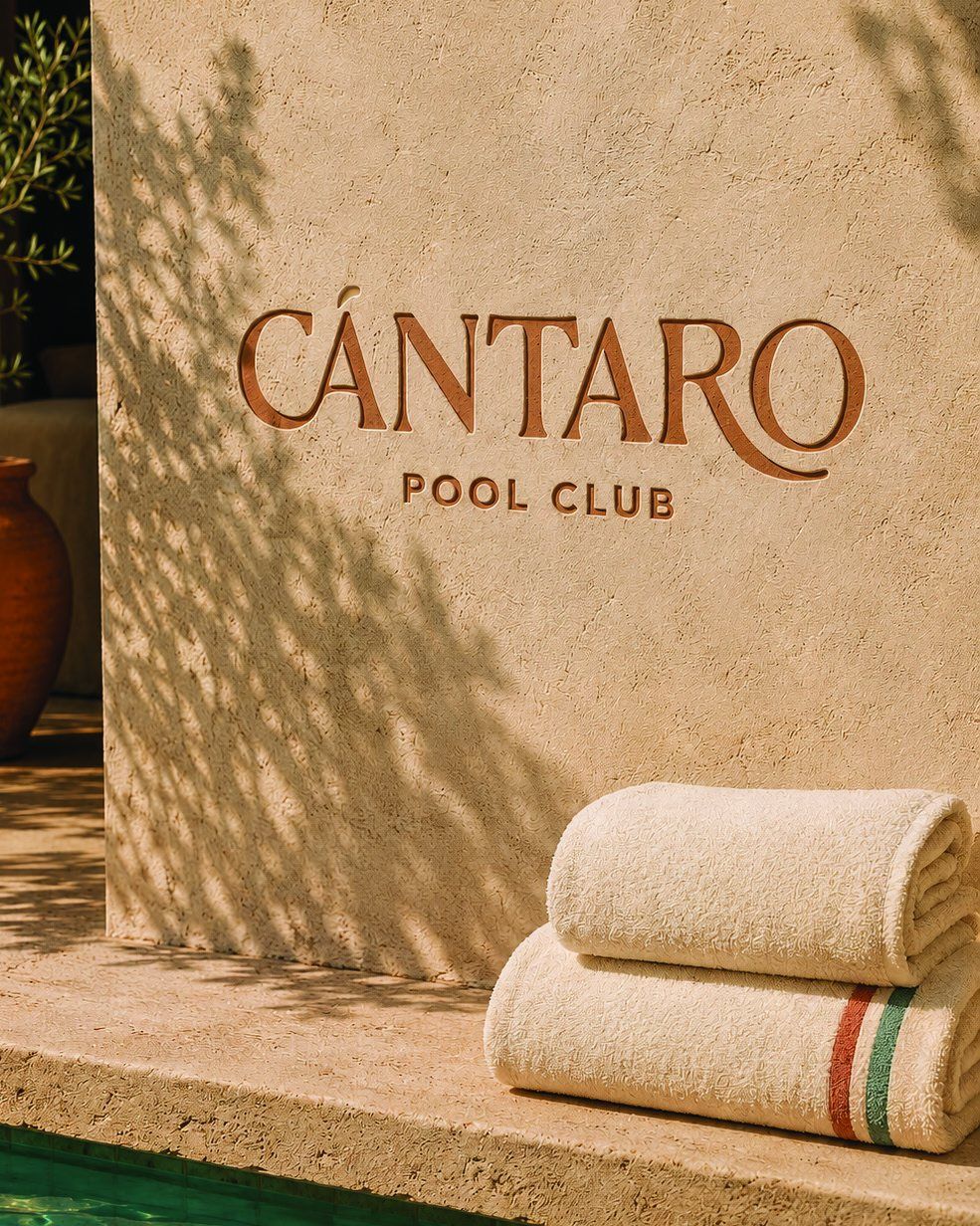 The Cántaro Pool Club wordmark cut into a stucco wall beside folded pool towels